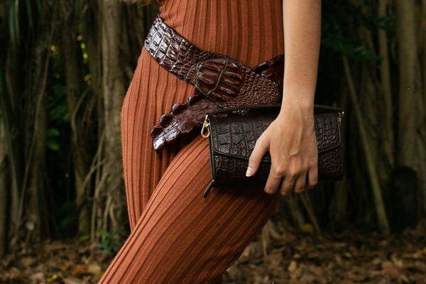 a girl wearing a crocodile skin backstrap belt while holding a crocodile skin clutch in brown